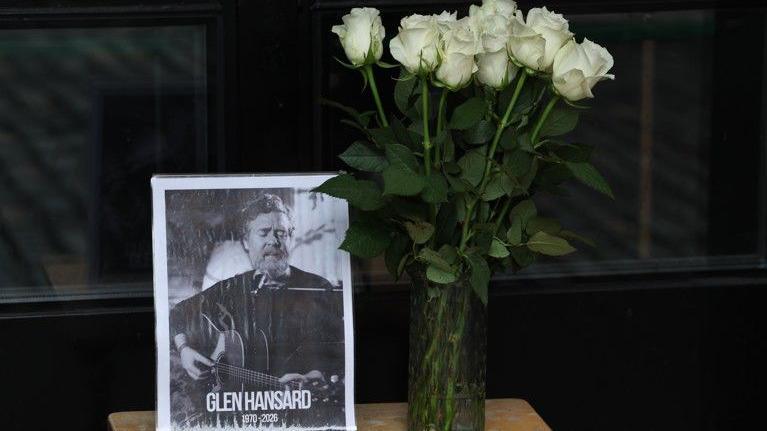 A black and white picture of Glen Hansard sits on a table with a vase of white roses beside it