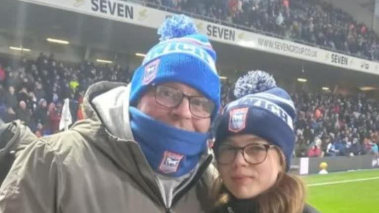 A man smiles at the camera while wearing a beanie hat and scarf around his mouth. He is also wearing glasses and a green coat. He stands with a young girl who is also wearing a beanie hat and brown glasses.