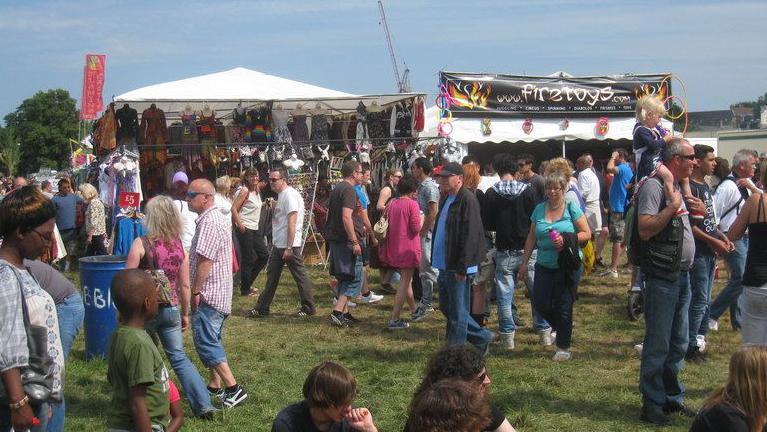 Crowds of men, women and children, walking on shaggy grass on a summer's day. Behind them are two stalls and in front are the heads of four people sitting on the grass, Strawberry Fair, Midsummer Common, 2011