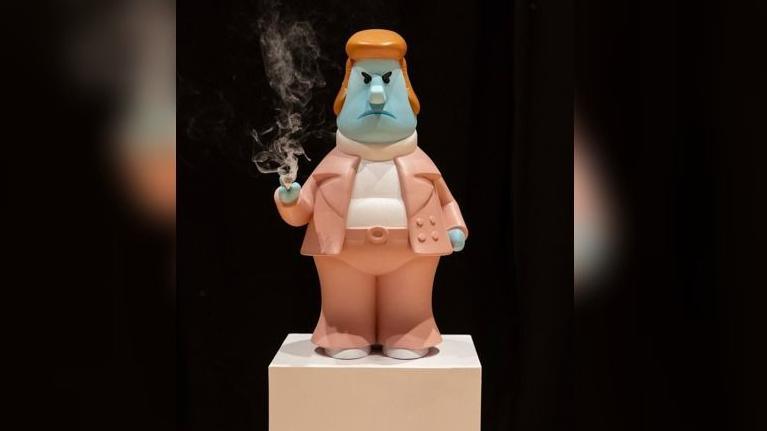A cartoon-style figure of a man with a blue face and orange hair is holding a smoking cigarette is on a cream coloured plinth. The figure is wearing a peach-coloured suit with a white polo neck top.