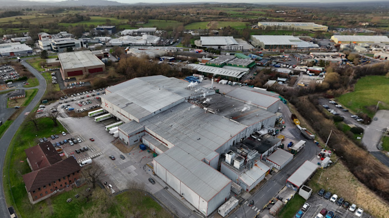 Birds eye view of Oscar Mayer factory at an industrial site in Wrexham. Car parks can be seen dotted around the large site, along with other warehouse buildings. Green hills can be seen in the far background.