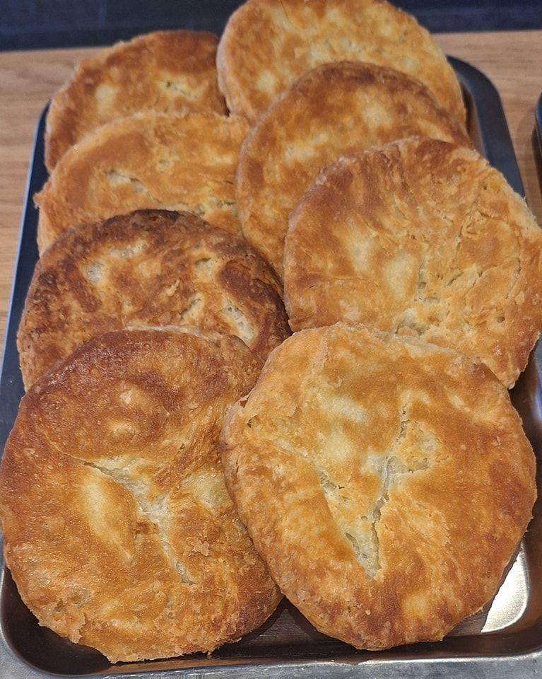 Six golden brown butteries - a round, flat food made of lard, butter and sugar - on top of a tray on a brown table.