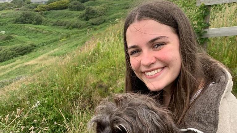 Maddy Lawrence is looking at the camera smiling with a dog on her lap. There is a sweeping green hillside in the background.