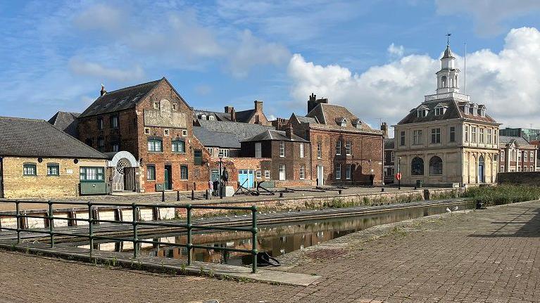 General view of King's Lynn's historic docks. Lots of ornate brick buildings connected by bridges with a body of water running through them. 