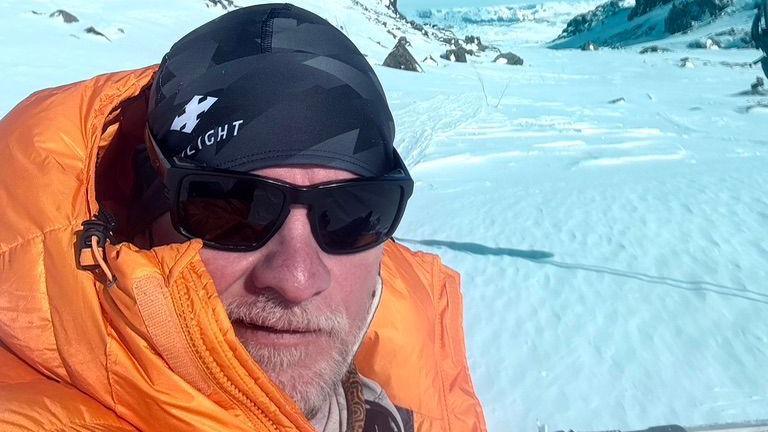 Ian Hughes, pictured in an icy valley with the mountains on either side behind him. He is wearing a black cap and sunglasses, and an orange hooded coat.
