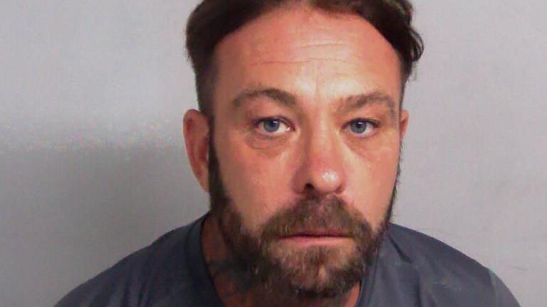 Phillip Curson in police custody. He is wearing a grey T-shirt and has brown hair and a beard.