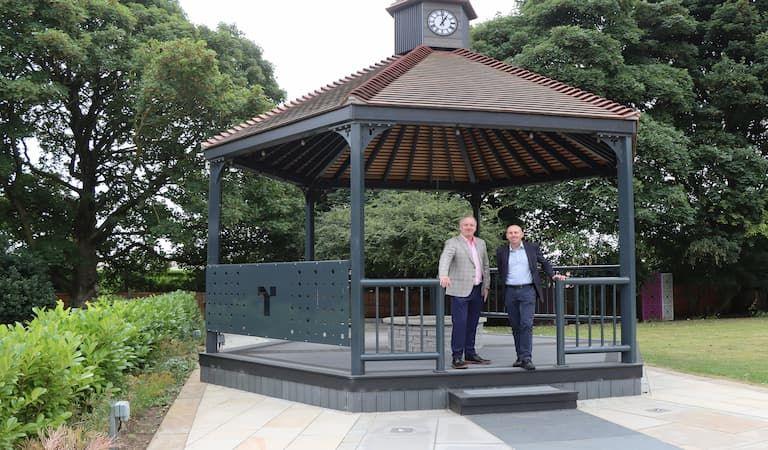 Mark Wall and Rob Waltham dressed in casual suits standing in a bandstand with tiled roof and clock and trees in background