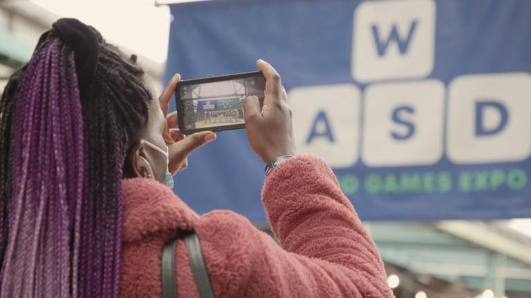 A view from the back of the head of a woman in a pink coat taking a picture on her phone of a large overhead banner of the WASD logo. 