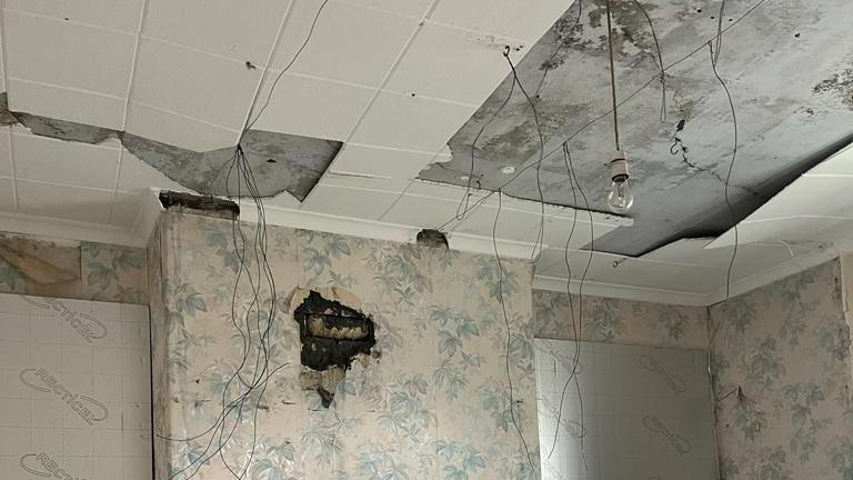 A poorly maintained ceiling with wires dangly freely and naked bulbs. Tiles are peeling off and mould appears to be growing on a wallpapered fireplace.