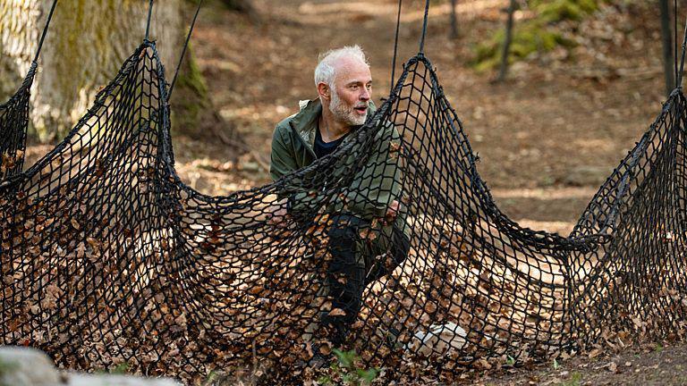 Mark Bonar is crouched on a forest floor and is about to get swept up in a net. His head is just visible about the thick, black netting.