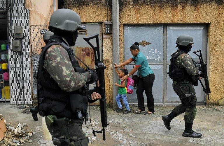 A mother and child walk past military police on patrol near the Vila Kennedy favela in Rio de Janeiro on February 23, 2018