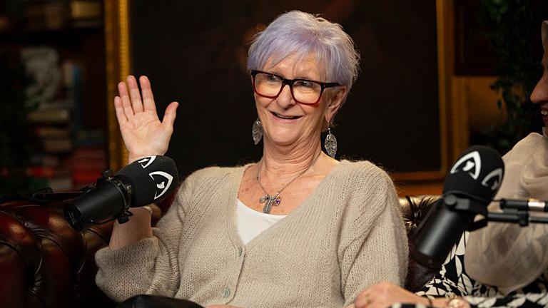 Linda sits on a brown leather sofa, waving to the other side of the room. A podcast microphone sits in front of her. She is wearing a cream cardigan, white T-shirt and leaf-shaped earrings. Her hair is purple-toned and she is wearing black-rimmed glasses.