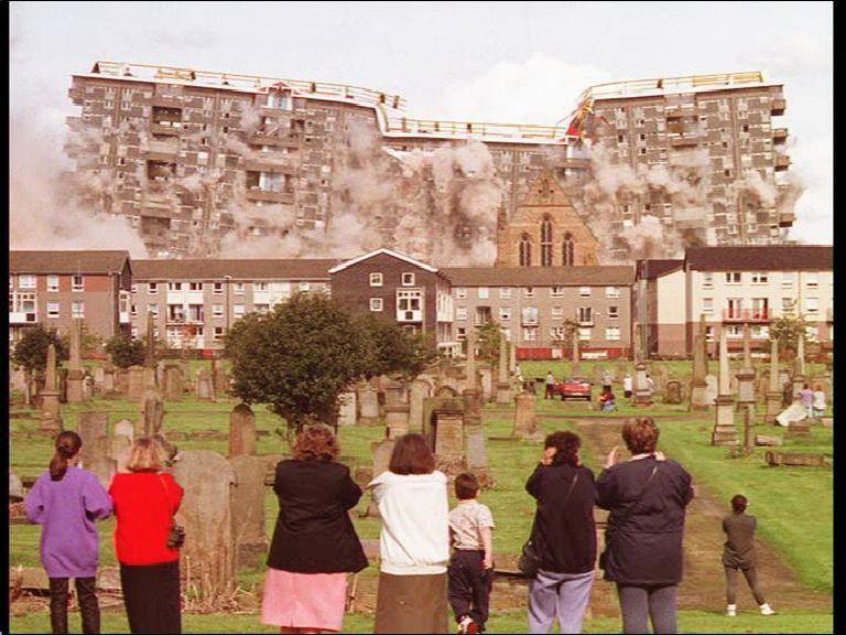 A head-on image of a row of high rise flats, mid-explosion. We look on from behind a row of people watching the whole thing. The flats are collapsing in in themselves from the middle out, plumes of dust going into the sky.