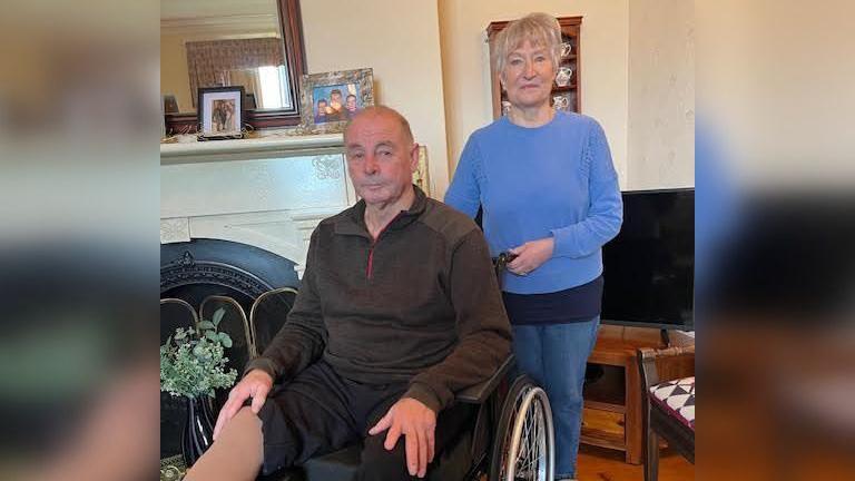 Alun mewn cadair olwyn yn ei gartref a'i wraig Anita yn sefyll y tu ôl iddo