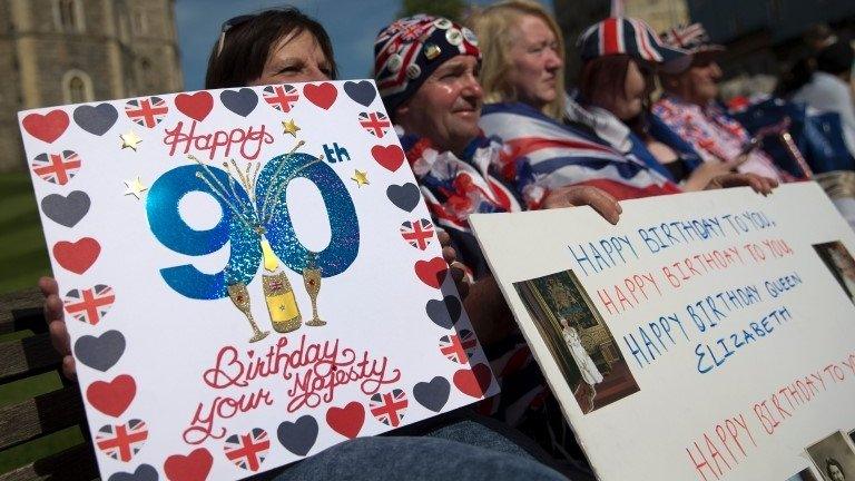 Royal well-wishers hold placards wishing Queen Elizabeth II a happy 90th birthday as they wait in hope of seeing her following her engagements in Windsor