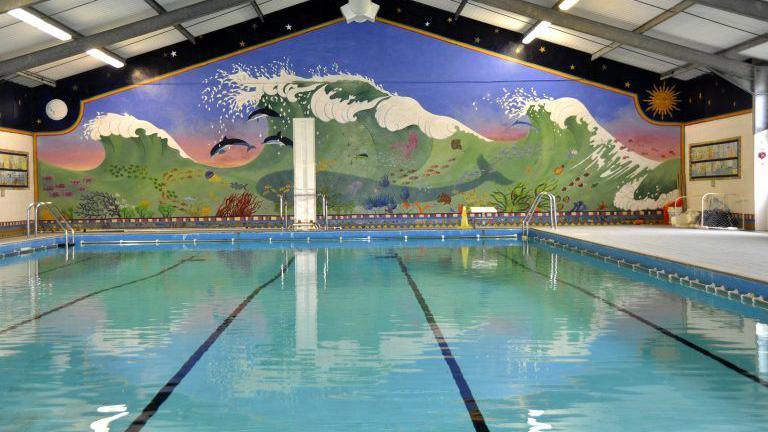 A swimming pool with blue tiles. On the wall behind it is a large mural of a blue/green wave. Inside the wave is a large whale, schools of fish and dolphins.