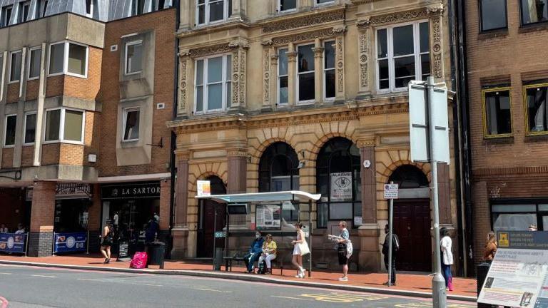 An old fashioned stone building on a high street behind a bus stop