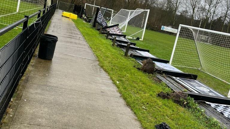 A concrete path running between two grass football pitches. On the right side, a fence has blown over and its stakes have come out of the ground. It lies flat on the grass, next to two white goals. A yellow bin is on its side in the distance and on the left side, a black metal fence stands firm. The ground is wet and the sky grey. Poplar trees are in the background. 