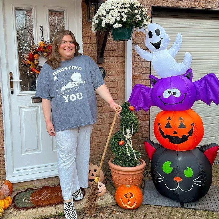 A woman with shoulder-length fair hair wearing whiote trousers and a grey t shirt with the words "ghosting you" on it. She is stood in front of a white front door with an orange, autumnal wreath hung on it. She is holding a broomstick and beside her are pumpkins, a potted plant with a skeleton attached, and a blow-up halloween decoration with a black cat, an orange pumpkin, a purple bat and a white ghost shape included.