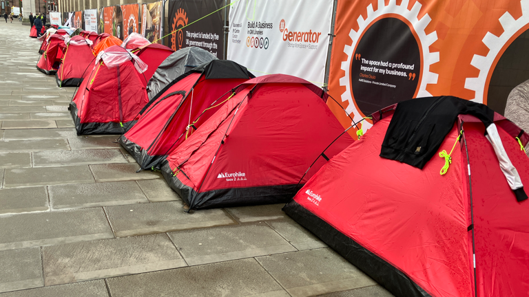 A line of red tents outside Manchester town hall, pitched on wet stone paving, under some fencing covered in advertising. 