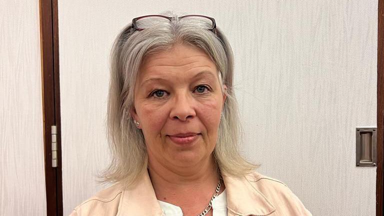 Sarah Jane Colclough wearing a cream jacket, with her glasses on her head. She has light grey to blonde hair down to her shoulders, a metal chain necklace around her neck and a cream jacket over a white top. She is in front of a grey coloured door.