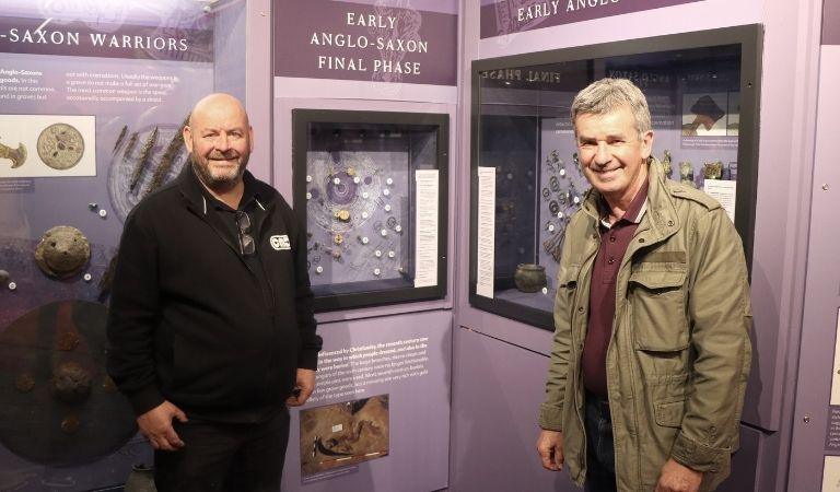 A bearded Craig Allison dressed in black standing next to John Davison, who is wearing a khaki jacket and a burgundy top. Some of the exhibits are in the background