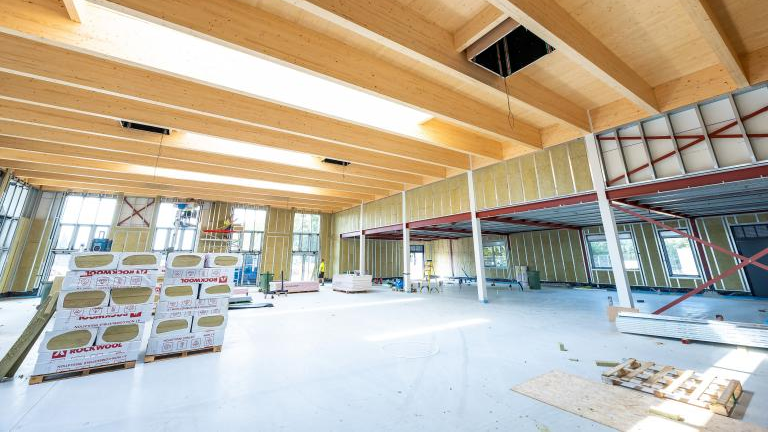 A picture showing the inside of the hall which is under construction. There is a pine-coloured roof and other building materials scattered throughout the hall.