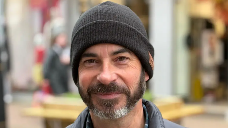 Mark Warren is smiling at the camera. He is wearing a black woolly beanie hat and has brown eyes and a brown and white beard. There are shops behind him.