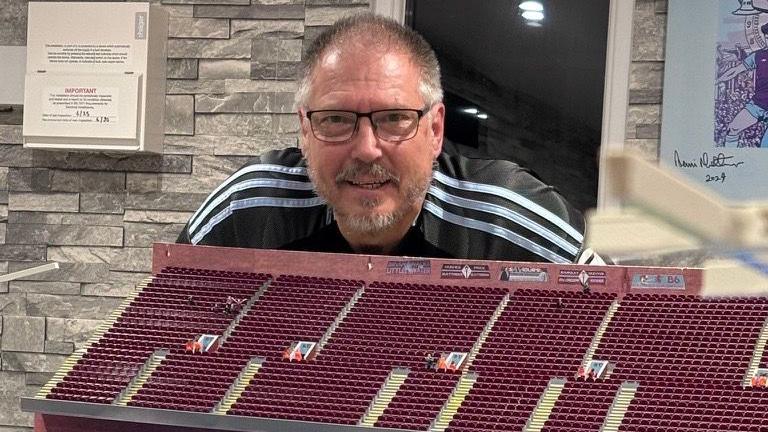 A man with grey hair, a beard and white glasses stands wearing a black and white football shirt behind part of a model of Villa Park football stadium. The model has 3D-printed plastic claret chairs in the stands. Behind the man is a brick wall with football memorabilia on it.
