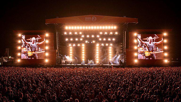 A view of the Radio 2 in the Park festival stage from far back. Thousands of people stand in front of the crowd watching Sting perform at night. The stage is lit up and has two large screens either side.