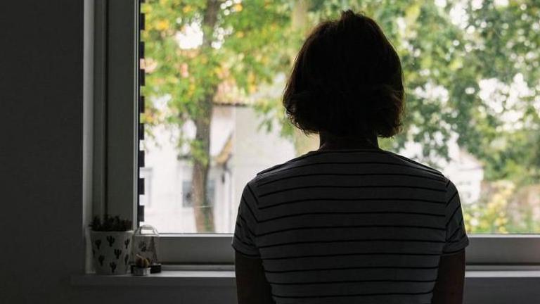 A woman sat in front of a window with her back to the camera. She has bobbed hair and is wearing a striped t-shirt