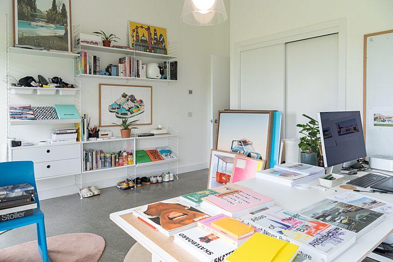 A modern, bright office all in white, with pops of colour coming from a multi-coloured trainer print, a bright blue chair, brightly-coloured book covers and a pink lightshade