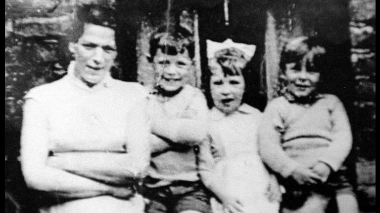 A black and white photo of a woman standing looking at the camera with her arms folded. Beside her are three children, also looking at the camera and smiling.