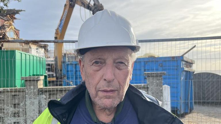A head and shoulders image of Bill Seal. He is wearing a hard hat and standing in front of a metal fence, behind which is a large blue tip and a yellow crane. He is looking directly into the camera.