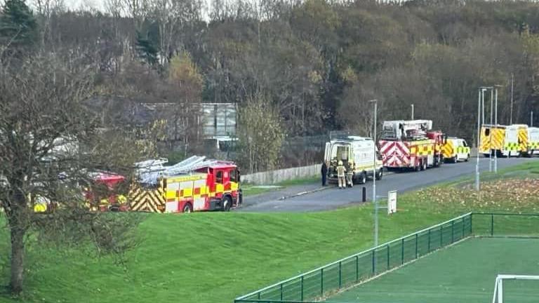 At least three fire engines can be seen parked in a street as well as ambulance and police vehicles. The road is bordered by trees on the far side and a grassy area in the foreground with a football pitch.