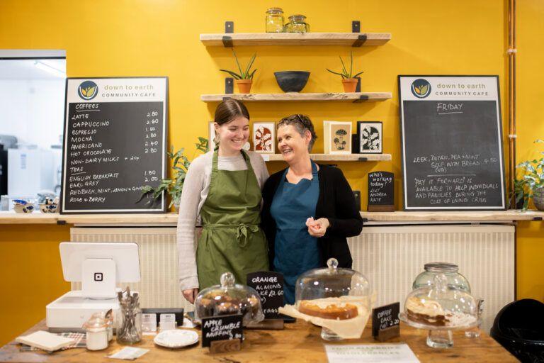 Two women wearing aprons smile at each other. In the background are menu boards showing coffee and food options. In the foreground is a table laden with cakes and a till.