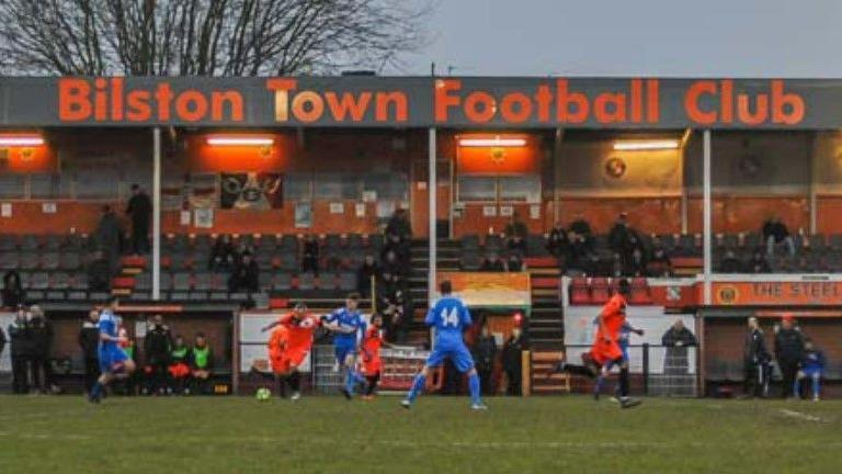 Players from two football teams in orange and blue play in a match, while spectators in a stand are visible behind. The top of the stand carries lettering spelling "Bilston Town Football Club" in orange.