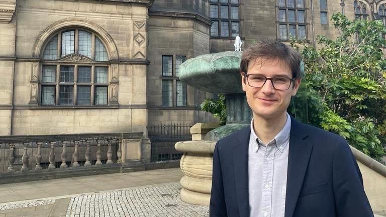 A man is standing outside Sheffield town hall, he has short brown hair, black rimmed glasses and is wearing a blue suit jacket and blue open necked shirt
