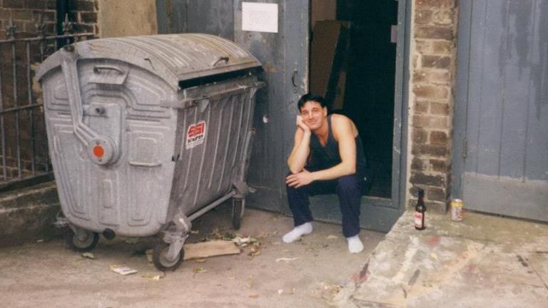 David Artus sitting in the doorway the back door of the theatre. He has short dark hair and is wearing a dark vest and trousers and white socks. There is a large metal bin on the left and a bottle of beer to the right.