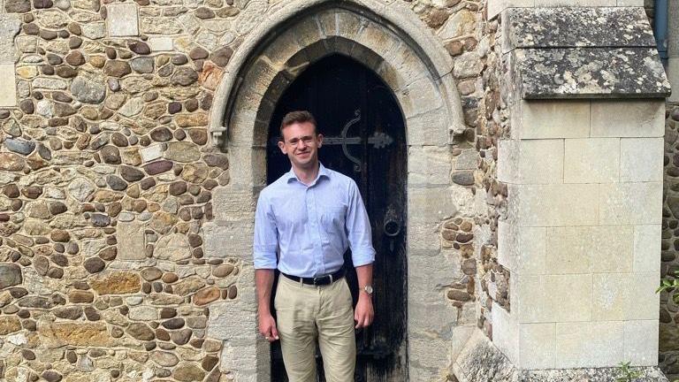 John Hager is standing in the doorway to a church. The building is made from stones and there an ornate black wooden door behind him and a stone arch above. He has short brown hair and is wearing glasses, a pale blue shirt and beige trousers