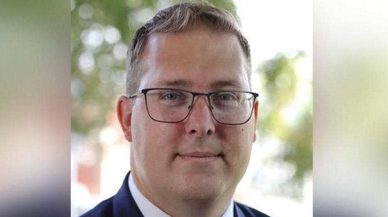 A head and shoulders photograph of Gareth Potter's face. He is looking directly at the camera, is wearing glasses and has a neutral expression. The blurred background appears to include trees with green foliage.