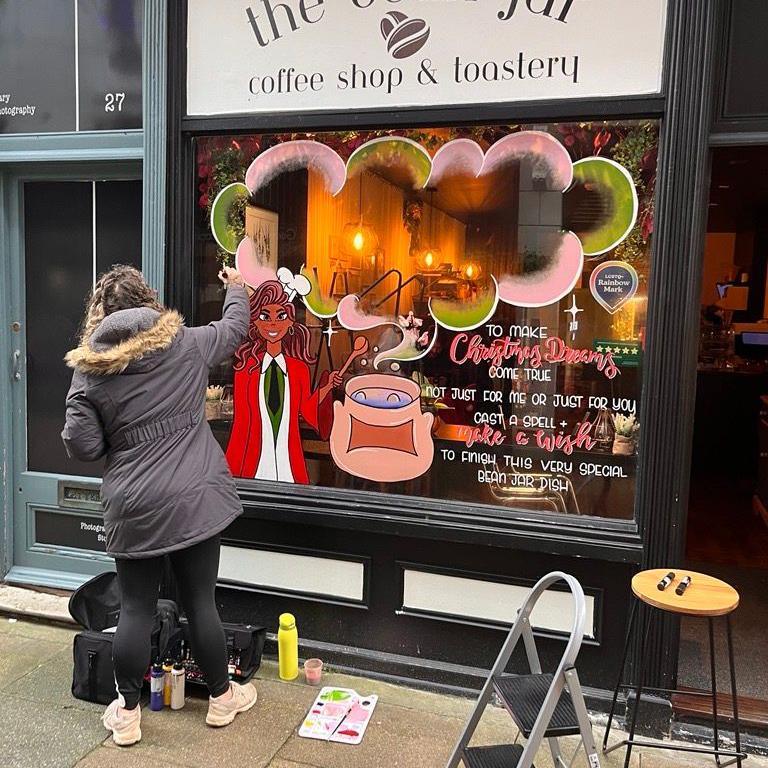 A woman in a grey coat with black leggings decorates a shop window with paints.