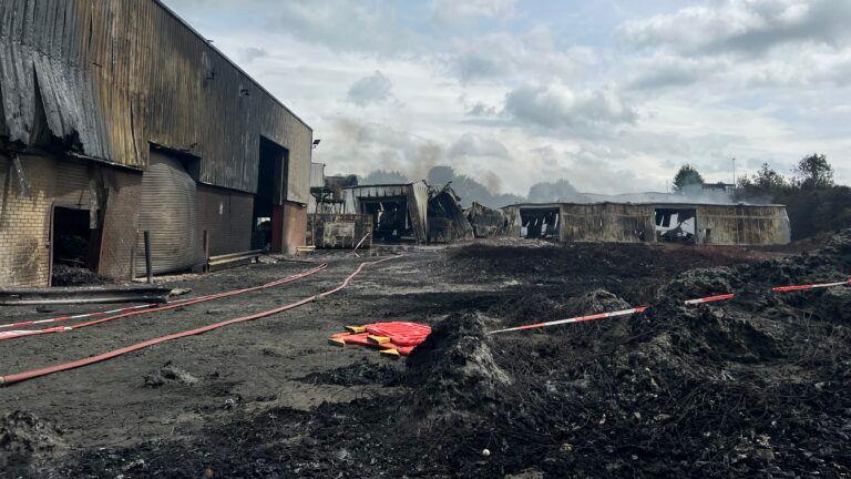 A burnt out building, showing burnt ground and burnt metal buildings, fire hoses and plastic barriers on the ground. 