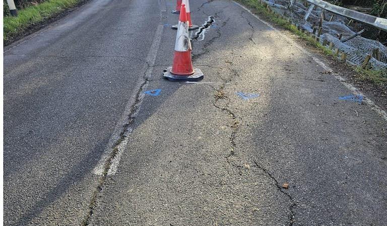 Image showing the damage to the road, There are a number of large cracks, as well as traffic cones and blue paint marks on the carriageway.