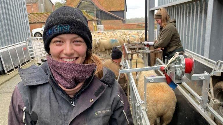 Hannah Murrell is wearing a black coat and black had that reads 'Ewe First' while looking at the camera and smiling. Behind here are some sheep being led through a metal framed walkway. 