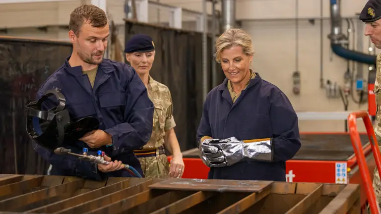 A woman wears a metallic glove while looking at a piece of machinery that is being explained by a man in blue overalls. Two soldiers stand either side.