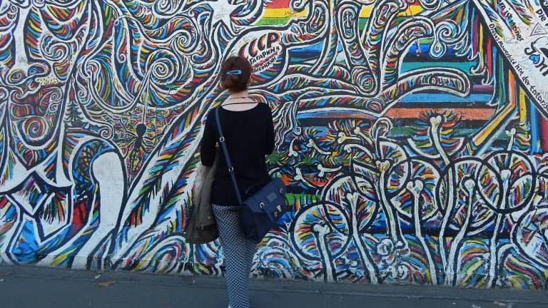 Dr Krysia Waldock photographed from behind looking at a wall of colourful graffiti. They are dressed in a navy top and trousers and has their hair pinned in a bun.