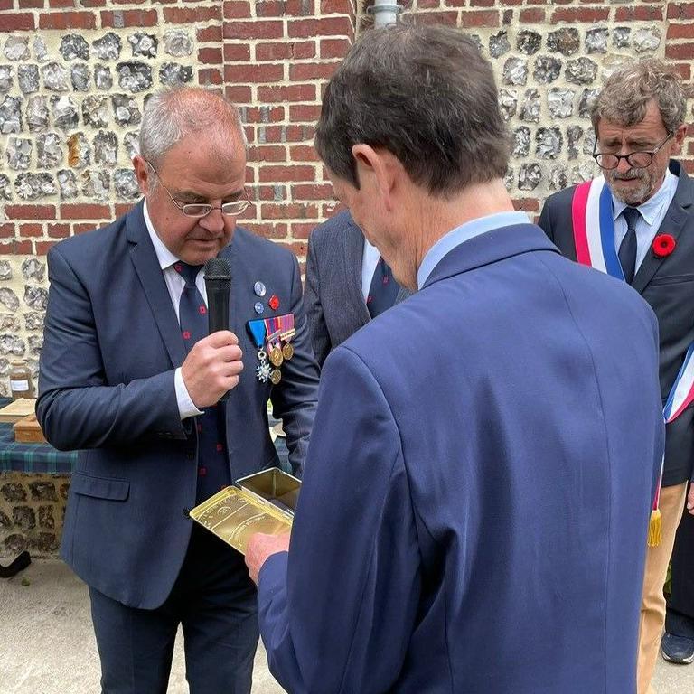 Mr Balfour, wearing a blue suit and facing away from the camera, looks down at a box as a smartly-dressed man wearing a blue suit and medals, speaks into a microphone. To the right, another man wears a poppy and a sash in the colours of the French tricolour flag.