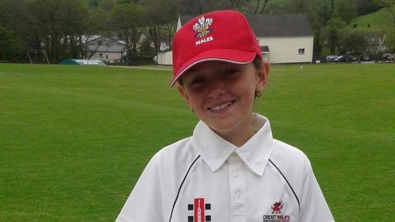 Libby wearing her white "Welsh Cricket/Criced Cymru" polo shirt and long white trousers  and a red Wales cap, pictured as a young teenager. She is standing in front of a white picket fence at a rural-looking cricket ground 