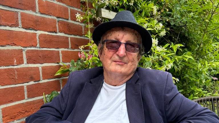 Andrew Lines smiles at the camera while sitting in a garden. He wears a black bowler hat, sunglasses, a navy suit jacket and white T-shirt.